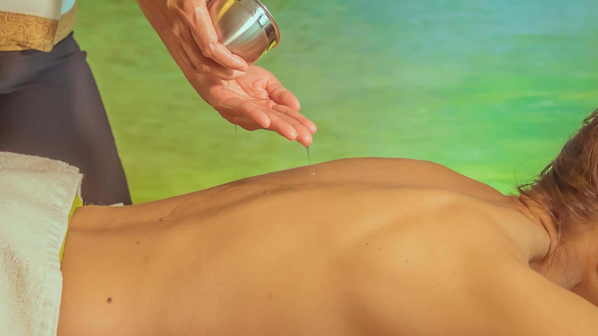 Zur Vorbereitung der Ölmassage tropft Massageöl aus einem Gefäß auf den Rücken der Frau, die auf der Massageliegt liegt.